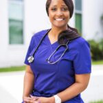 Portrait of a smiling female nurse in purple scrubs with a stethoscope, sitting outdoors.