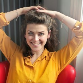 Cheerful woman sitting indoors, smiling brightly with hands behind head, wearing a yellow shirt.
